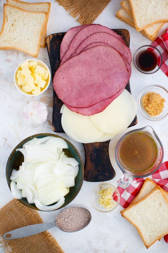 Overhead shot of French dip grilled cheese ingredients including roast beef, provolone, bread, butter, onions, and au jus seasoning arranged in bowls on a table.