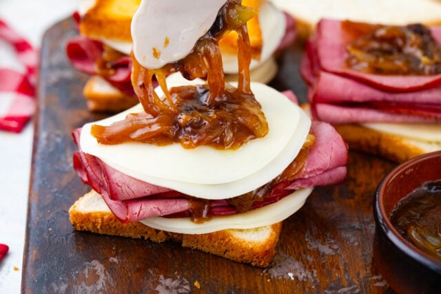 Assembling the French dip grilled cheese sandwich with layers of provolone, roast beef, and caramelized onions.