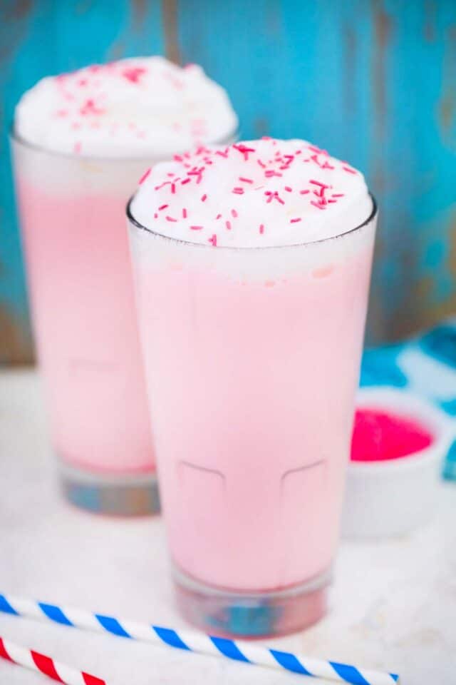 two tall glasses of pink angel milk topped with whipped cream and pink sprinkles